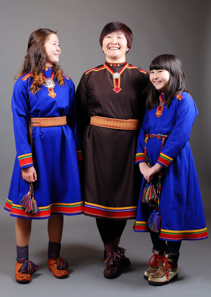 Marina sammen med de to døtrene sine. Alle kledt i Lyngenkofte. Jentene har blå kofter. Marina har mørkebrun kofte. Alle smiler.