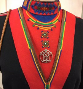 Nærbilde av koftebryst. Rød og svart, med pynt i grønt, gult og blått. Kråkesølvapplikasjoner og sølvbrosje.