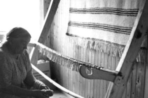 Eldre kvinne sitter ved siden av veven og nøster tråder. Grenen er klippet løs nederst og henger ned fra veven.