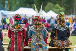 Tre damer i tradisjonelle drakter sitter på benk med ryggen til på Riddu.
